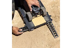 A person's hands positioning a small RC crawler onto an Axial 1/10 scale flatbed trailer with deployed loading ramps on a dirt surface.