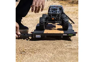 A person's hand adjusting the ramp on an Axial 1/10 scale flat bed vehicle trailer with an RC crawler loaded on top, sitting on a dirt surface.