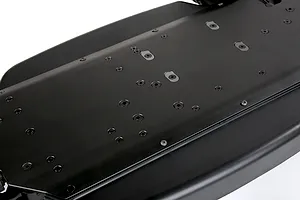 A close-up, angled view of a black aluminium RC car main chassis showing the underside with numerous countersunk screw holes.