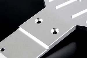 A close-up view of a silver aluminium RC front skid plate showing machined mounting holes and recessed channels against a black background.