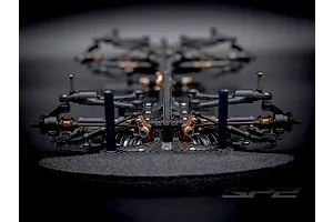 Low angle front view of the RC Maker SP2 EP touring car chassis showing carbon fiber, metal, and plastic components on a dark background.