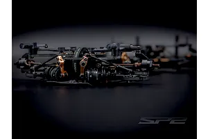Close-up studio shot of the RC Maker SP2 EP touring car front suspension, showing black and bronze components and drive belts on a dark background.