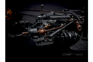 Close-up studio shot of the RC Maker SP2 EP RC touring car front suspension, showing carbon fiber, black plastic, and bronze-colored metallic parts.