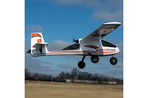 HobbyZone AeroScout S 2 RC airplane flying outdoors against a blue sky with clouds and a distant horizon.