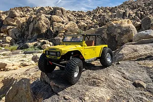 A yellow RC rock crawler with a roll cage traversing a rocky outdoor landscape in daylight.