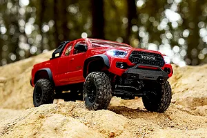 FMS FCX24M Toyota Tacoma red 1/24 scale RC rock crawler driving on a sandy dirt hill in an outdoor forest setting.