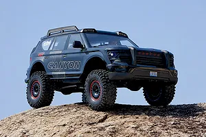 FMS Canyon RS RC rock crawler driving on a rocky outdoor surface against a clear blue sky background.
