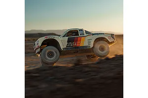 ARRMA Mojave 6S RC short course truck driving through dirt in a desert landscape at sunset, kicking up dust and airborne over jumps.