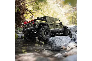 Green Axial RC rock crawler truck driving through a shallow rocky stream outdoors.