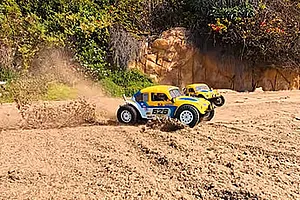Two yellow and blue Volkswagen Beetle RC buggies driving fast and kicking up dust on an outdoor dirt track.