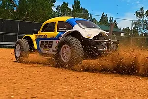 RC Volkswagen Beetle buggy driving and kicking up dirt on an outdoor dirt track with a chain-link fence background.