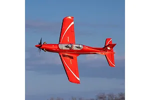 A red Pilatus PC-21 RC airplane in flight against a blue sky, viewed from above showing its wings and cockpit.