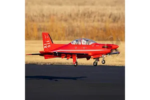 E-Flite Pilatus PC-21 RC plane in flight with red livery over a runway and blurred field background.