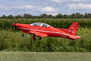 Red RC Pilatus PC-21 airplane in flight over a grassy field with landing gear extended.