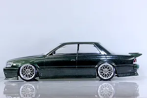 Side profile view of a painted green metallic RC drift car body shell mounted on a chassis with custom wheels against a white studio background.