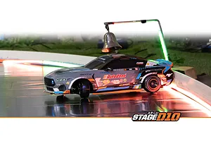 HPI Stage-D10 Ford Mustang drift RC car driving on an indoor track illuminated by red and green lights, passing under a suspended bell.