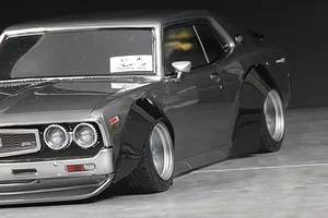 Close-up of a grey RC car body installed with wide black overfenders over deep-dish wheels against a dark background.