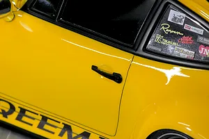 Close-up of a yellow RC car body showing the door handle, side window, and decals on the rear quarter glass.