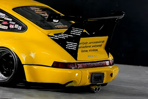 Close-up rear angle of a yellow RC body shell featuring a large black spoiler, custom decals, wide fenders, and twin exhaust pipes.