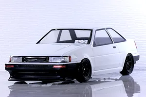 Studio photo of a white Toyota AE86 Corolla Levin 2-door RC car body with black wheels and front bumper against a light brick wall background.