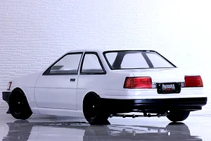 Rear-three-quarter view of a white Toyota AE86 Corolla Levin RC car body with black wheels on a reflective surface.