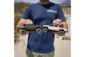 A person holding two small RC pickup trucks, one green and one beige, in an outdoor desert environment.