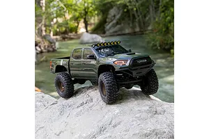 Green Axial Toyota Tacoma RC rock crawler truck parked on a large grey boulder near a stream in an outdoor setting.