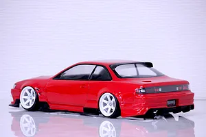 Rear quarter view of a red RC drift car body styled after an S14 Silvia with white wheels and black aero parts on a reflective white surface.