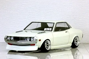 White Toyota Celica 1600GT RC drift car body shell mounted on a chassis with custom wheels and a textured white brick background.
