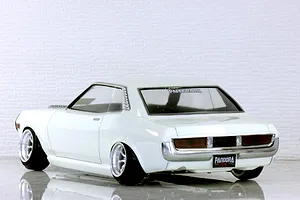 Rear-three-quarter view of a white RC drift car body on a reflective surface with white brick wall background.