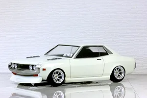 White Toyota Celica 1600GT RC body shell mounted on a chassis with custom wheels against a textured white brick background.