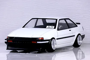 Front three-quarter view of a white Toyota AE86 Sprinter Trueno 2-door RC drift car body shell on a reflective surface with a white tiled wall background.