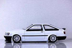 Side profile view of a white Toyota AE86 Corolla Levin RC drift car body on a reflective surface against a white brick wall background.
