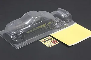Unpainted clear polycarbonate RC car body shell with a mask sheet and sticker sheet on a dark surface.