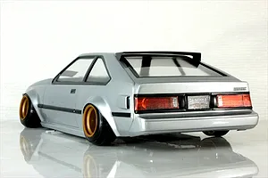 Rear three-quarter view of a painted silver RC drift car body on a reflective white surface with gold wheels and black trim.