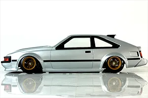 Side profile view of a silver Toyota Celica RC drift car body with gold mesh wheels on a white reflective surface.