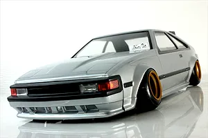 Studio product photo of a silver Toyota Celica XX A60 RC drift car body shell mounted on a chassis with gold-rimmed wheels.