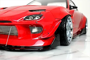 Close-up front quarter view of a red RC drift car body with wide body kit, aftermarket front bumper, and chrome wheels on a marble surface.
