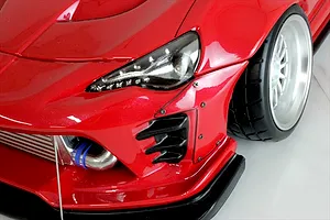 Close-up of the front left section of a red RC car body, showing the headlight, front wheel, bumper, and intercooler.