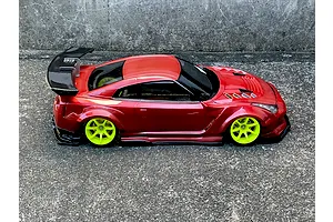 Side profile of a red metallic RC drift car body shell with a large rear wing and bright green wheels on asphalt.