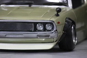 Close-up studio view of the front corner of a gold RC body shell showing headlights, grille, fender flare, and bumper.