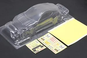 Unpainted clear polycarbonate RC car body shell with a rear wing, window masks, and sticker sheets on a dark surface.