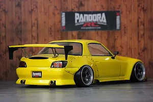 Rear-three-quarter view of a yellow Honda S2000 RC drift car with widebody fenders and large rear wing against a wooden background.