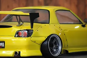 Close-up rear quarter view of a yellow radio-controlled drift car body featuring a large black wing, wide fender flares, and detailed wheels.