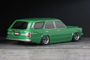 Rear three-quarter view of a green RC Mazda RX-3 Savanna Sports Wagon body mounted on a chassis with custom mesh wheels against a dark studio background.