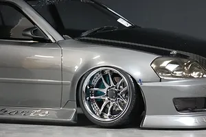 Close-up of a silver RC drift car body showing the front wheel, chrome multi-spoke rim, and black hood.
