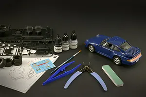 An assembled blue Porsche 911 model car alongside plastic parts sprues, tools, paint bottles, and instruction sheet on a dark background.