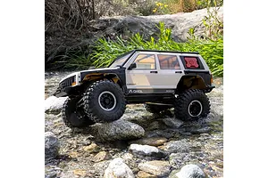 Axial SCX10 III Jeep Cherokee XJ RC rock crawler driving over rocks and through a shallow stream in an outdoor setting.