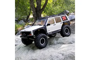 Axial SCX10 III Jeep Cherokee XJ white RC rock crawler driving outdoors on rocky terrain among trees.