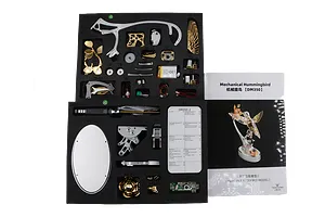 Overhead studio view of the Teching DM350 Mechanical Hummingbird parts laid out in black foam trays next to its instruction manual.